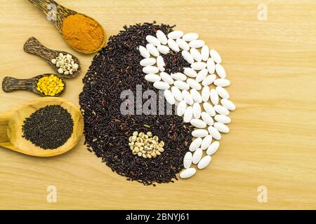 Schwarzer Reis und weiße Pille bilden ein Yin Yang Symbol und Kurkuma-Pulver, Hirse, Sojabohne, Basilikumsamen in Holzlöffel auf braunem Holz zeigen BL Stockfoto