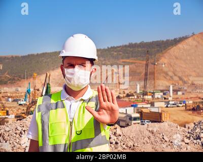 Ingenieur in medizinischer Maske zeigt Stoppschild gegenüber der Baustelle. Stockfoto