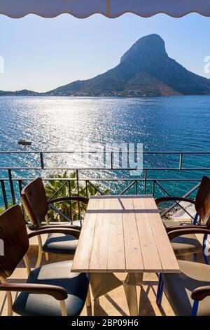 Meerblick auf die Insel Telendos über einen Tisch und Stühle auf einem Balkon im Ferienort Masouri, Kalymnos, Dodekanes, Griechenland Stockfoto