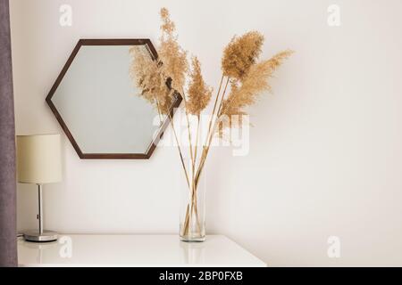 Trockenes Schilf in einer transparenten Glasvase, die neben dem Holzspiegel steht. Stockfoto