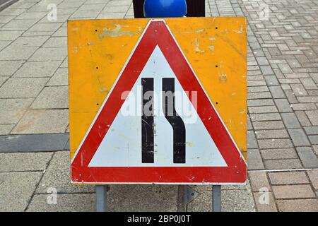 Dreieckige rot-weiße Straßenschild UK Road Engerging auf der rechten Seite, auf gelber Tafel. Ramponiert aussehende Schild auf asphaltierter Straße, städtische Umgebung. Stockfoto