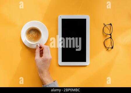Hand des Mannes mit einer Tasse Kaffee, Tablet mit leerem Bildschirm und Brille auf gelbem Hintergrund, Stockfoto