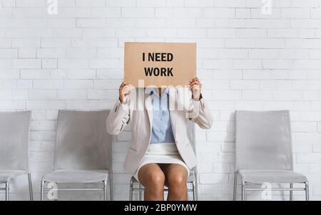 Schwarze Frau in Business-tragen versteckt hinter Zeichen mit Inschrift Ich BRAUCHE ARBEIT, während warten auf Arbeit Interview im Büro Stockfoto