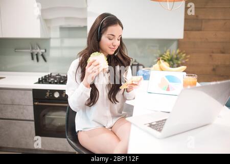 Schöne schwarzhaarige Frau arbeitet von zu Hause aus. Ein Mitarbeiter sitzt in der Küche und hat viel Arbeit auf einem Laptop und Tablet und hat Videokonferenzen Stockfoto