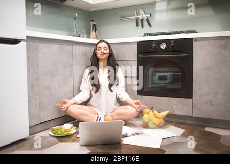 Schöne schwarzhaarige Frau arbeitet von zu Hause aus. Ein weibliches Model sitzt in einer Yoga-Pose auf dem Küchenboden. Das Ende einer ausgewogenen und ruhigen Arbeit. Fühlt sich voll an Stockfoto
