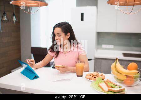 Schöne schwarzhaarige Frau arbeitet von zu Hause aus. Ein Mitarbeiter sitzt in der Küche und hat viel Arbeit auf einem Laptop und Tablet und hat Videokonferenzen Stockfoto