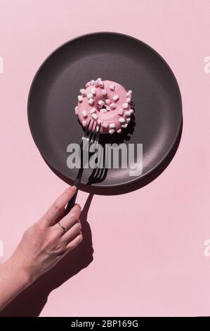 Hellrosa Delicious donat mit weißen Marshmallows an der dunkelgrauen Keramikplatte auf dem rosigen Hintergrund. Hand der Frau mit Ring mit Sommersprossen ist Stockfoto