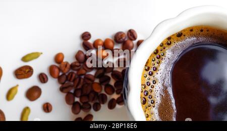 Flaches Lay. Banner. Schwarzer Kaffee mit Kaffeeschaum in einer weißen Tasse. Im Hintergrund sind Kaffeebohnen und Kardamom-Körner auf einer weißen glänzenden Oberfläche. Stockfoto