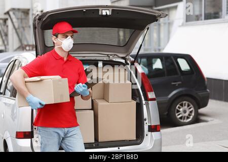 Kurier in Uniform, Schutzmaske und Handschuhe steht in der Nähe Kofferraum des Autos mit Paketen Stockfoto