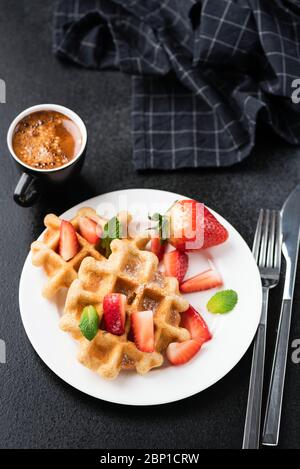 Süße knusprige Waffeln mit Erdbeeren und einer Tasse Espresso auf schwarzem Hintergrund. Vertikale Ausrichtung. Stillleben von Waffeln mit Beeren und Kaffee Stockfoto