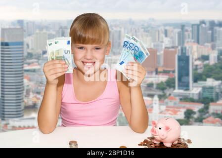 Kleines fröhliches Mädchen, das mit Münzen und rosa Sparschwein auf dem Schreibtisch sitzt und europäische Währung über dem Hintergrund der Stadt in Händen hält Stockfoto