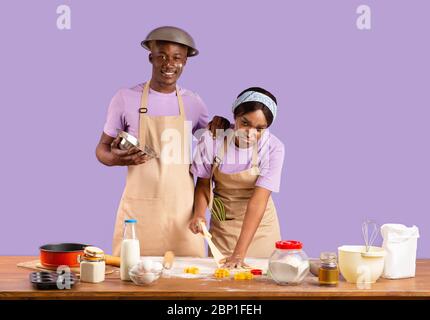 Happy guy und erschöpft Mädchen mit Mehl von Kopf bis Fuß bedeckt beim Kochen auf violettem Hintergrund Stockfoto