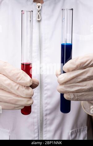 Zwei Fläschchen mit blauer und roter Flüssigkeit, in Handschuhen vor dem Hintergrund eines Arztes. Entwicklung von Impfstoffen und Medikamenten. Medizinische und wissenschaftliche Bio Stockfoto
