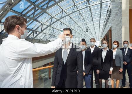 Arzt trägt schützende chirurgische Maske mit Infrarot-Stirnthermometer (Thermometer Pistole), um Körpertemperatur auf Virussymptome des Geschäfts zu überprüfen Stockfoto