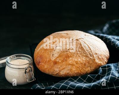 Weizenrunder Sauerteig Brot und Sauerteigstarter im Glasgefäß. Seitenansicht von köstlichen hausgemachten Sauerteig Brot auf schwarzem Hintergrund mit Kopierraum. Hausgemachtes Sauerteig Brot machen Konzept. Stockfoto