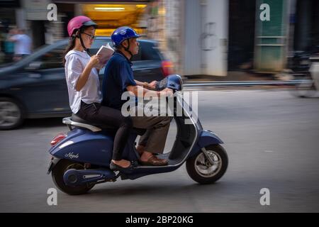 Hanoi, Vietnam - 30. April 2018: Mädchen liest auf einem Motorrad, während ihr Vater mit beweglichem Hintergrund fährt. Stockfoto