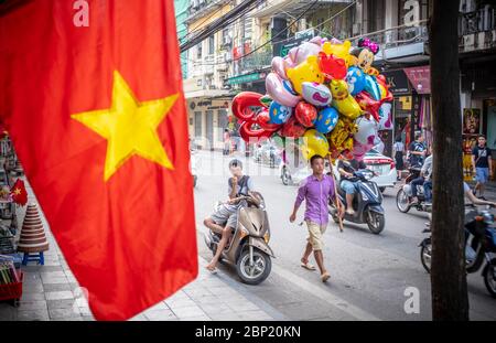 Hanoi, Vietnam - 30. April 2018: Straßenszene mit einem lokalen Händler, der einen Haufen bunter Ballons und die vietnamesische Nationalflagge im Vorschiff trägt Stockfoto