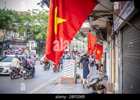 Hanoi, Vietnam - 30. April 2018: Straßenszene mit Einheimischen, die ihre Fahrräder oder Motorräder auf einer belebten Straße mit vietnamesischer Nationalflagge befreien Stockfoto