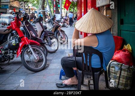 Hanoi, Vietnam - 30. April 2018: Straßenszene mit einem Mann, der einen Strohhut trägt und von hinten gesehen Motorräder auf dem Bürgersteig parkt Stockfoto