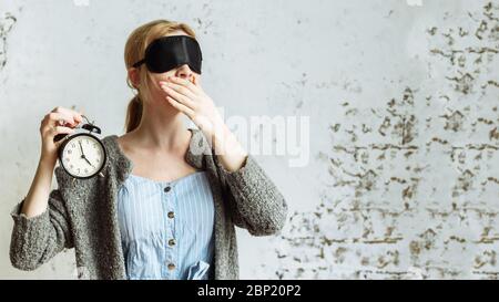 Eine Frau in einer Schlafmaske hält einen Wecker in der Hand. Sie ist müde und faul am Morgen. Das Konzept der Schlafprobleme. Stockfoto