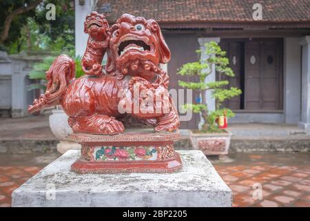 Traditioneller asiatischer Löwe aus rotem Porzellan, aufgenommen in einem Stadtpark mit Bonsai-Bäumen im Hintergrund, Hanoi, Vietnam Stockfoto