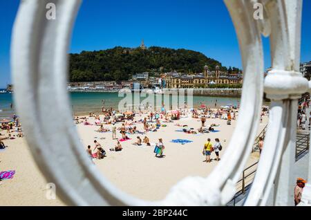 Donostia, Gipuzkoa, Baskenland, Spanien - 12. Juli 2019 : Blick durch das ikonische Geländer des Strandes von La Concha. Sonnenanbeter und Urgull Hügel im Hintergrund Stockfoto