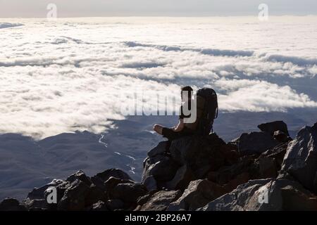 Ein Bergsteiger, der hoch auf dem Mount Ruapehu sitzt und über die Wolken schaut, Tongariro National Park, Neuseeland Stockfoto