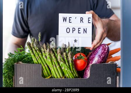 Ende der Sperrung der Quarantäne. Mann hält Leuchtkasten mit Grußtext-Nachricht Wir sind offen und frische Gemüse und Gemüse-Box. Einladender Lebensmittelgeschäft cli Stockfoto