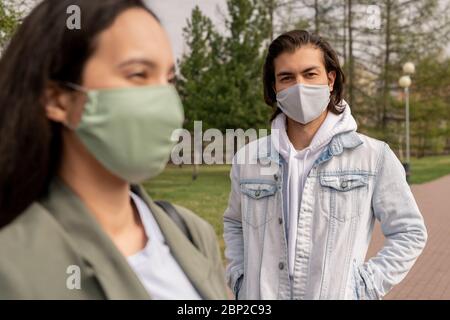 Schöner junger Mann in Schutzmaske und Freizeitkleidung, der Sie anschaut, während er mit seiner Freundin am Sommertag im öffentlichen Park spazieren geht Stockfoto