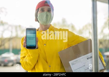 Junge Kurierin in Arbeitskleidung und Maske zeigt Transaktionsdaten auf dem Smartphone-Bildschirm, während auf den Kunden vor der Tür warten Stockfoto