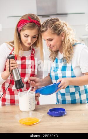 Blonde Mutter hilft Tochter beim Backen in der Küche mit karierten Schürzen. Stockfoto