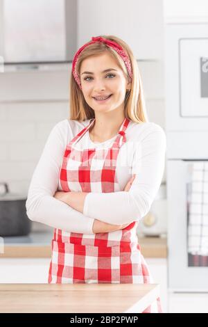 Teenager-Mädchen trägt rot karierte Schürze mit Hosenträger steht mit verschränkten Armen auf der Brust in der Küche. Stockfoto