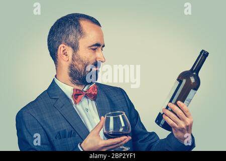 Ein Mann mit einem Glas Wein hält, schaut auf eine Flasche, lesen Sie die Informationen darüber. Gemischte Rasse bärtige Modell isoliert auf grün-gelbem Hintergrund Stockfoto
