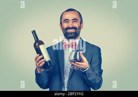 Ein gutaussehender, fröhlicher, lächelnder Geschäftsmann mit einer Flasche Wein und Glas im Studio. Gemischtes Modell mit Bartmuster auf grün-gelbem Hintergrund isoliert. Position Stockfoto