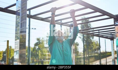 Morgendliches Workout. Starker und gesunder bärtiger Senior in Sportbekleidung, der beim Training im Stadion an der horizontalen Stange Pull-Ups macht Stockfoto
