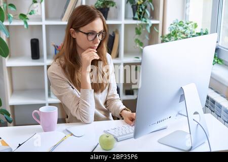 Online-Arbeit. Junge konzentrierte Geschäftsfrau, attraktive weibliche Mitarbeiterin in Brille am Schreibtisch sitzend und am Computer zu Hause arbeitend Stockfoto