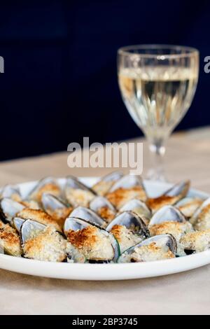 Miesmuscheln mit Parmesan, Brotkrume und Kräuterbutter auf einem Teller serviert. Ein Glas Weißwein. Hintergrund der Speisen. Mediterrane Küche. Konzept für eine Stockfoto