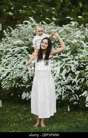 Junge Frau in einem langen weißen Kleid mit einem süßen kleinen Jungen in Hemd und Shorts zu Fuß in den grünen blühenden Garten am Sommertag Stockfoto