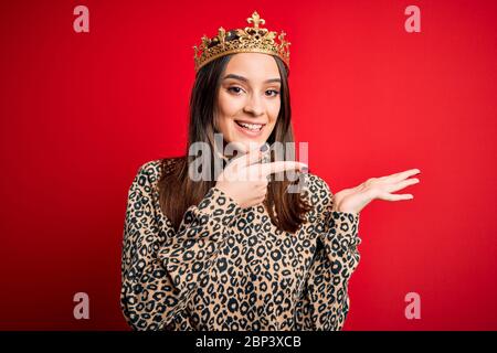 Junge schöne Brünette Frau trägt goldene Krone Königin über isoliert roten Hintergrund erstaunt und lächelnd an die Kamera, während mit der Hand und präsentieren Stockfoto