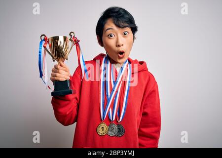 Junge schöne asiatische Mädchen Gewinner mit Trophäe trägt Medaillen auf weißem Hintergrund erschrocken in Schock mit einer Überraschung Gesicht, Angst und aufgeregt mit fe Stockfoto