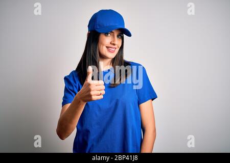 Junge Lieferfrau mit blauen Augen trägt Mütze über blauem Hintergrund tun glücklich Daumen nach oben Geste mit der Hand. Abzeichnen von Ausdruck, der sich ansieht Stockfoto