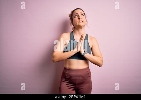 Junge schöne blonde Sportlerin, die Sport macht und Sportswear über rosafarbenem Hintergrund trägt, bettelt und betet mit den Händen zusammen mit dem Hope Expression On Stockfoto