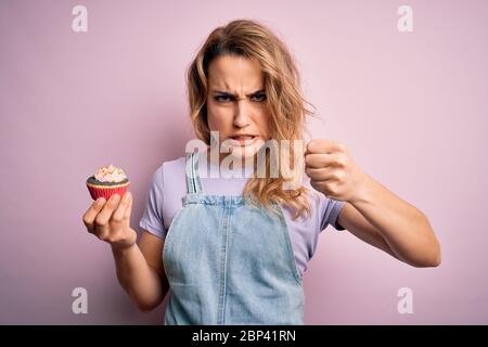 Junge schöne blonde Frau eatimg Schokolade Cupcake über isoliert rosa Hintergrund verärgert und frustriert schreien vor Wut, verrückt und schreien mit Stockfoto