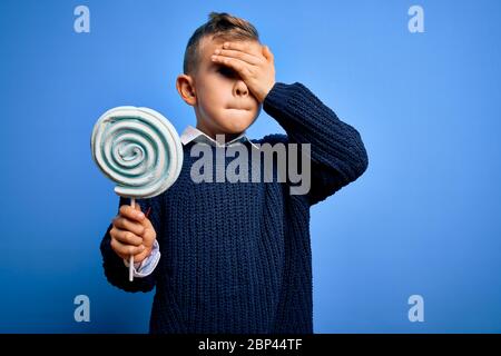Junge kleine kaukasische Kind essen süße Süßigkeiten Lutscher über blau isoliert Hintergrund mit Hand auf Kopf gestresst, schockiert mit Scham und Überraschung Gesicht, Stockfoto