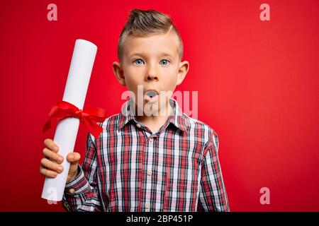 Junge kleine kaukasische Schüler Kind hält Schulabschluss über roten isolierten Hintergrund erschrocken mit einem Überraschungsgesicht, Angst und aufgeregt mit fe Stockfoto