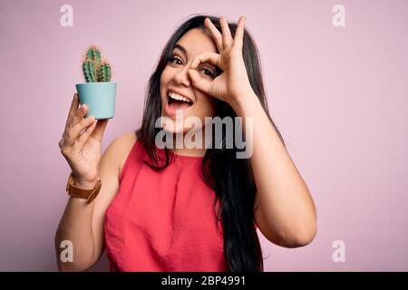 Junge Brünette Frau hält saftige Kaktus Pflanze über rosa Hintergrund mit glücklichem Gesicht lächelnd tun ok Zeichen mit Hand auf Auge durch Finger suchen Stockfoto