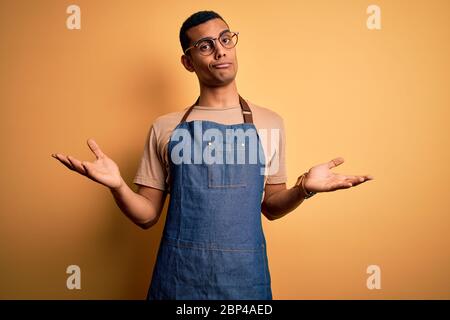 Junger schöner afroamerikanischer Ladenbesitzer, der Schürze über gelbem Hintergrund trägt, ahnungslos und verworren Ausdruck mit erhobenen Armen und Händen. Zweifel Stockfoto