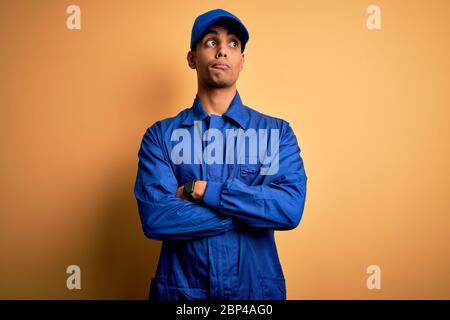 Junger afroamerikanischer Mechaniker Mann, der blaue Uniform und Kappe über gelbem Hintergrund trägt, lächelnd zur Seite schauend und wegstarrend denkend. Stockfoto