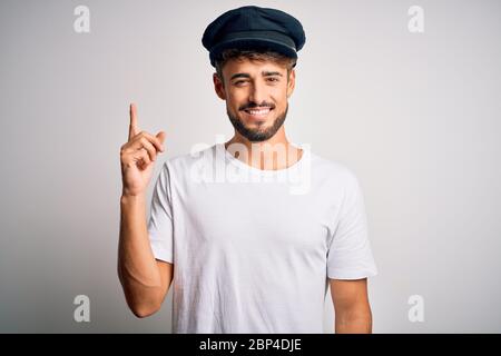 Junger Fahrer Mann mit Bart trägt Hut über isoliert weißen Hintergrund mit einem großen Lächeln auf dem Gesicht stehen, mit Hand und Finger zur Seite lo Stockfoto