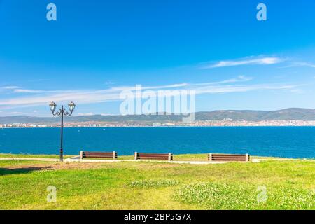 Drei Bänke am sonnigen Strand. Schöne Skyline Blick aus leerem Park mit gepflasterten Fußweg am Meer. Stadt und Berg in der Ferne b Stockfoto